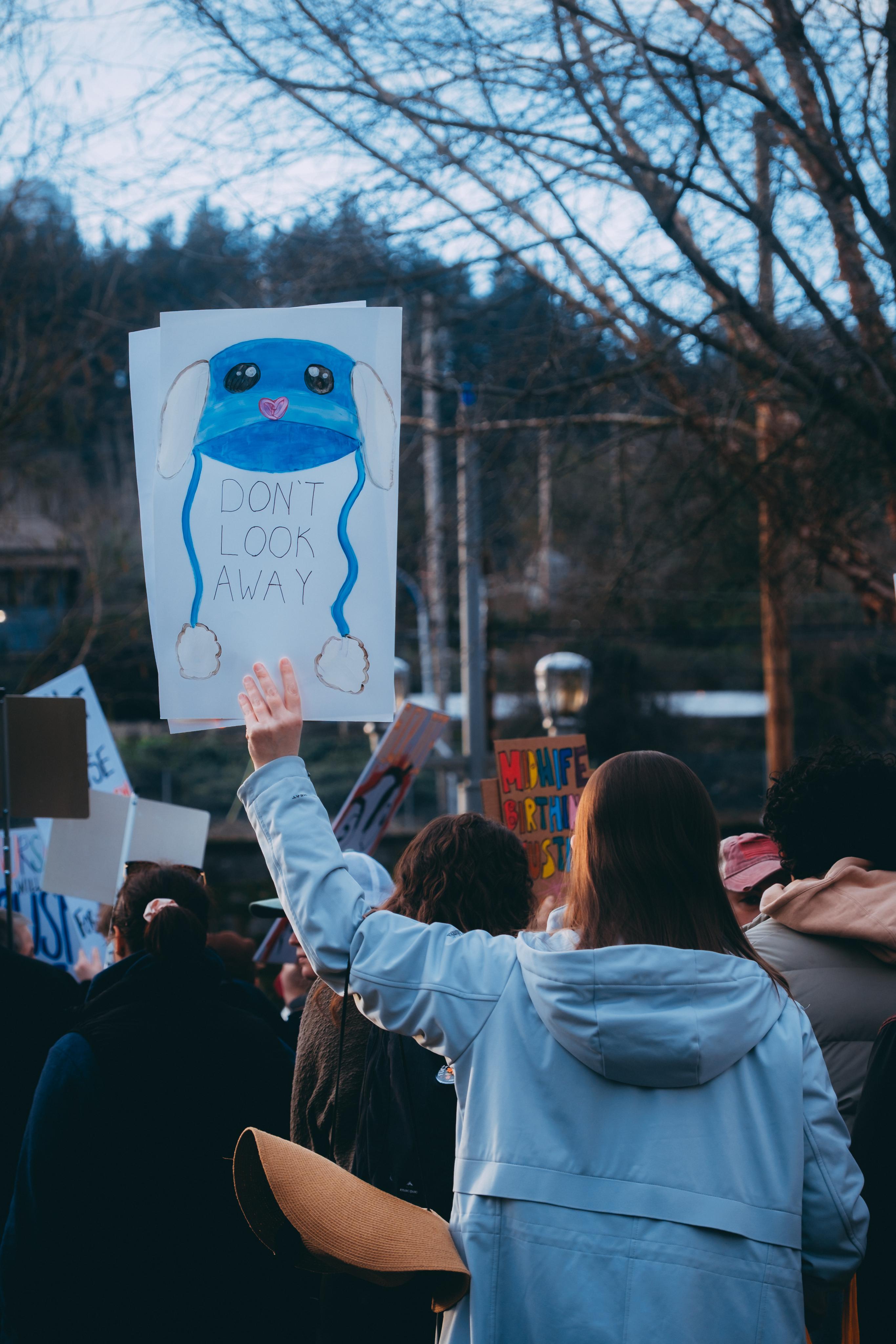 a person holding a sign that says don't look away with a drawing of a blue toque
